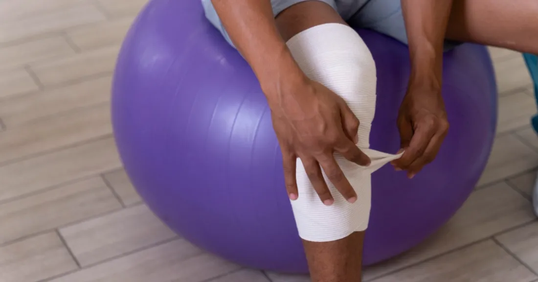 Patient sitting on a stability ball while wrapping a bandaged knee as part of total knee replacement rehabilitation.