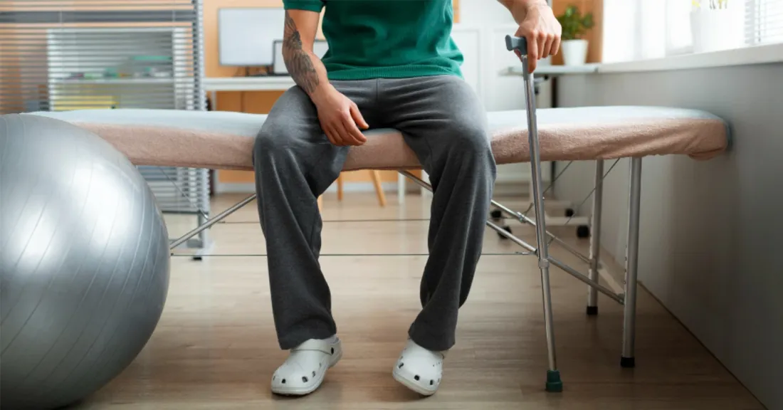 Patient with knee injury sitting on a rehabilitation treatment table using a cane, preparing for knee rehabilitation and recovery.