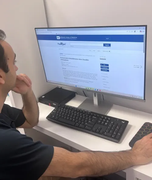 Physiotherapist studying PubMed article on shoulder replacement post-op rehab, showing the evidence gap our platform addresses.